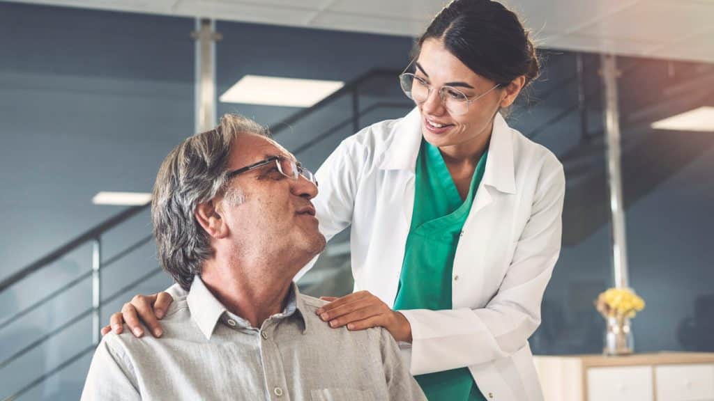 Older man wearing glasses and a gray button up shirt is being comforted by a female doctor while in a doctors office. 