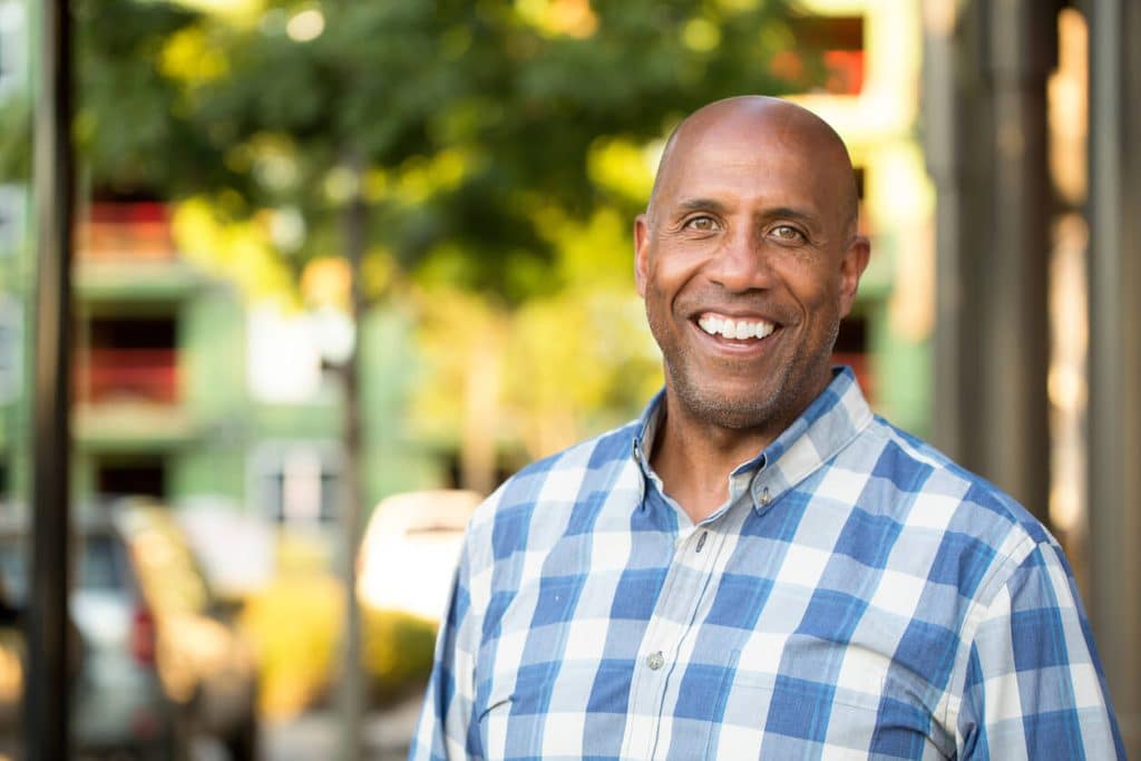 An older man standing outdoors on a sunny day wearing a white and blue shirt and smiling.