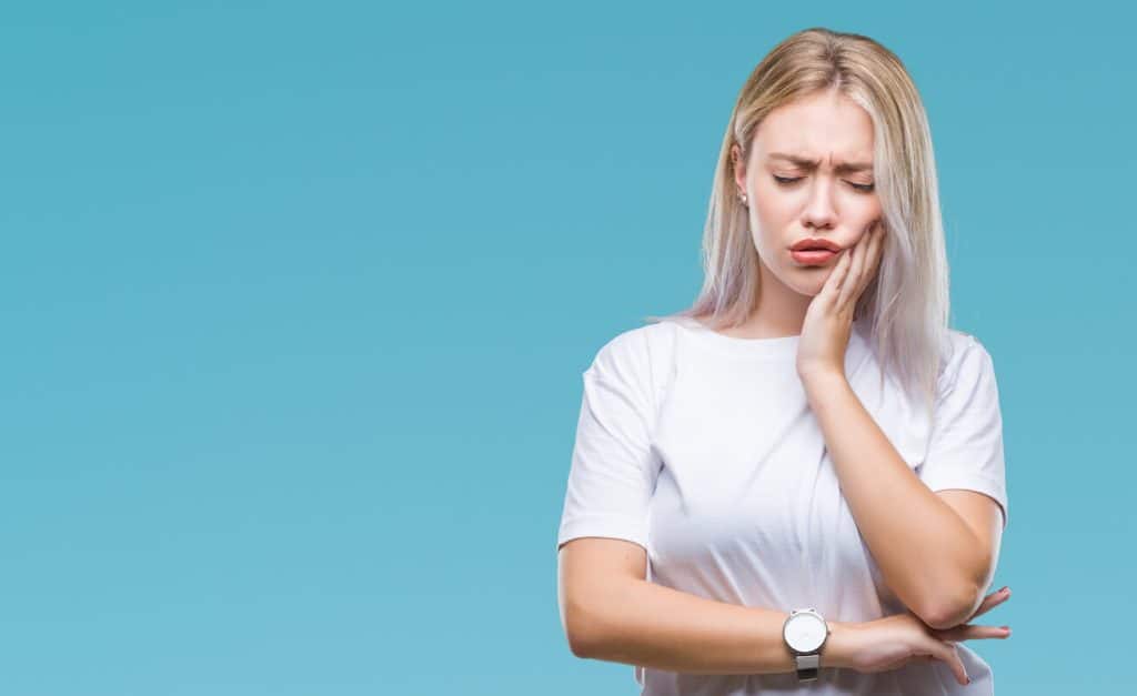 Blonde woman wearing a watch and a short sleeved shirt holding her mouth in pain in front of a blue background.